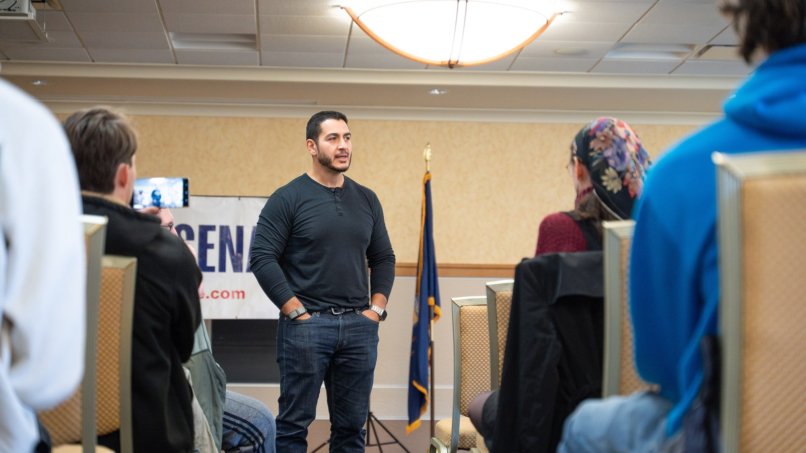 Abdul El-Sayed, wearing a dark sweater, stands and speaks to seated voters in front of a campaign banner at Michigan Technological University.